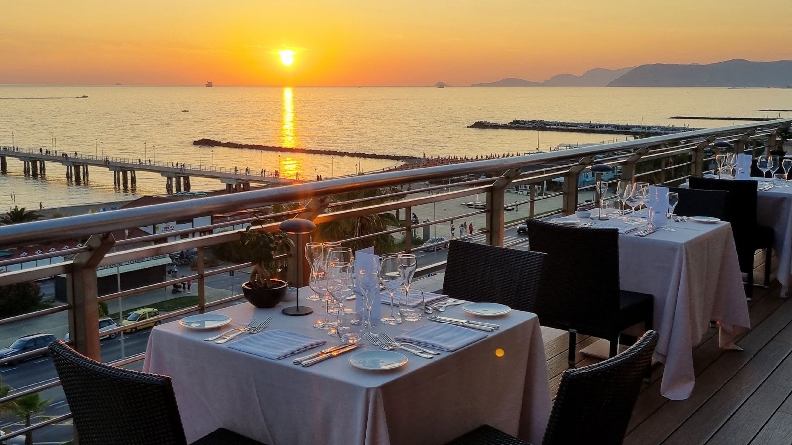 Comedor con mesas dispuestas para una comida, con vista a un cuerpo de agua y un atardecer al fondo.