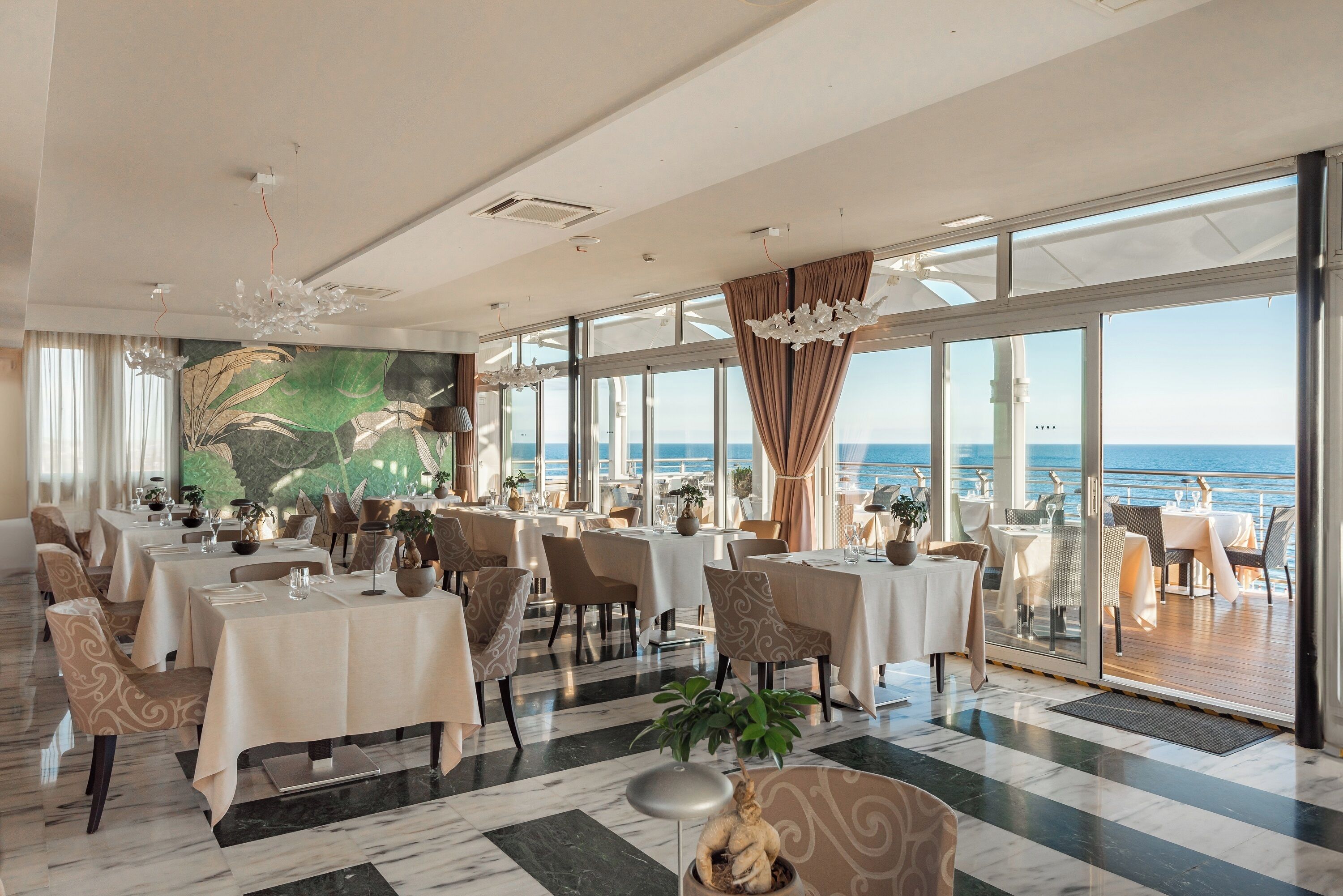 Un'ampia sala da pranzo con tavoli apparecchiati per gli ospiti, grandi finestre che offrono vista sul mare e un murale sulla parete.
