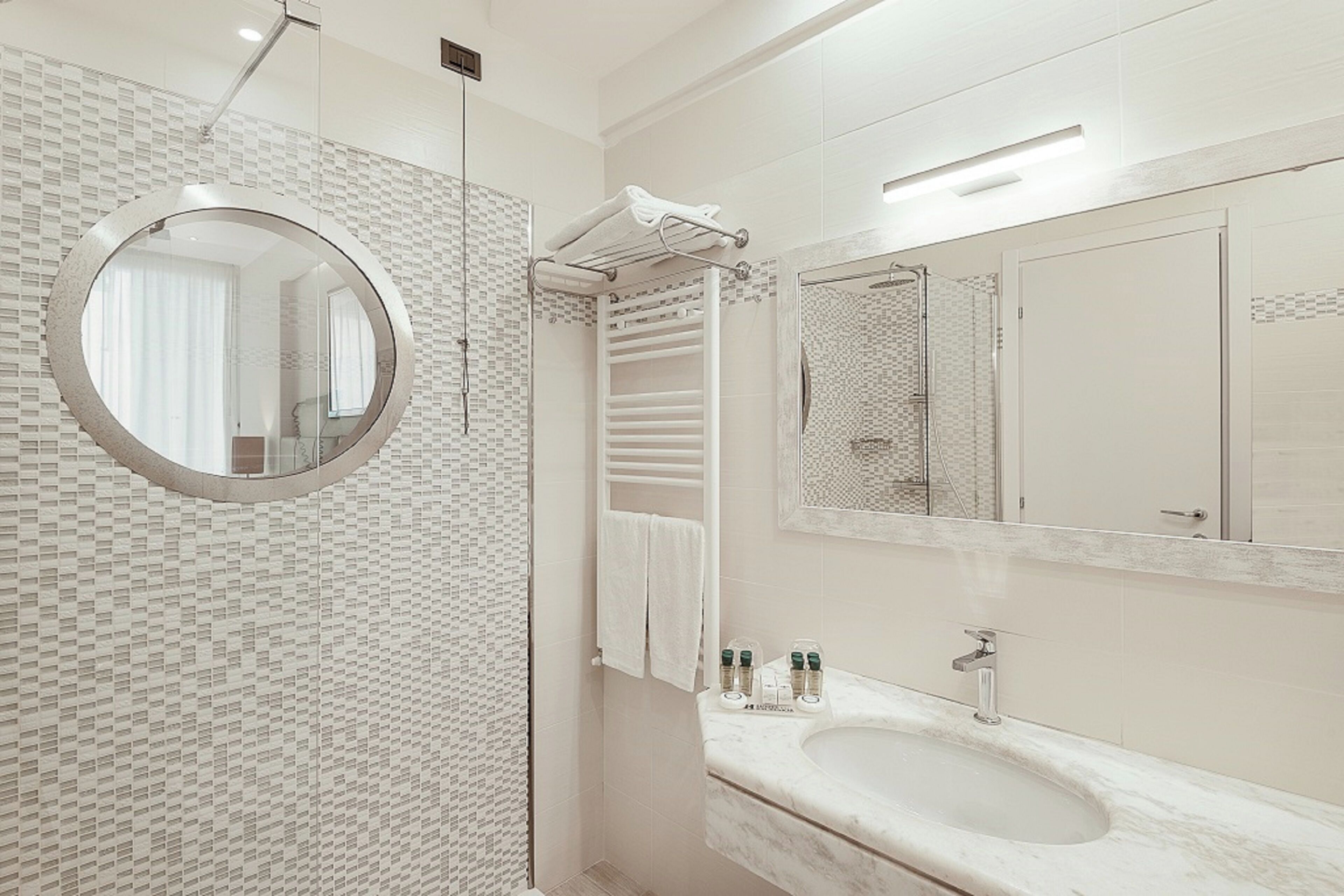 A modern bathroom with a round mirror, a glass shower enclosure, a marble sink, and a towel rack.