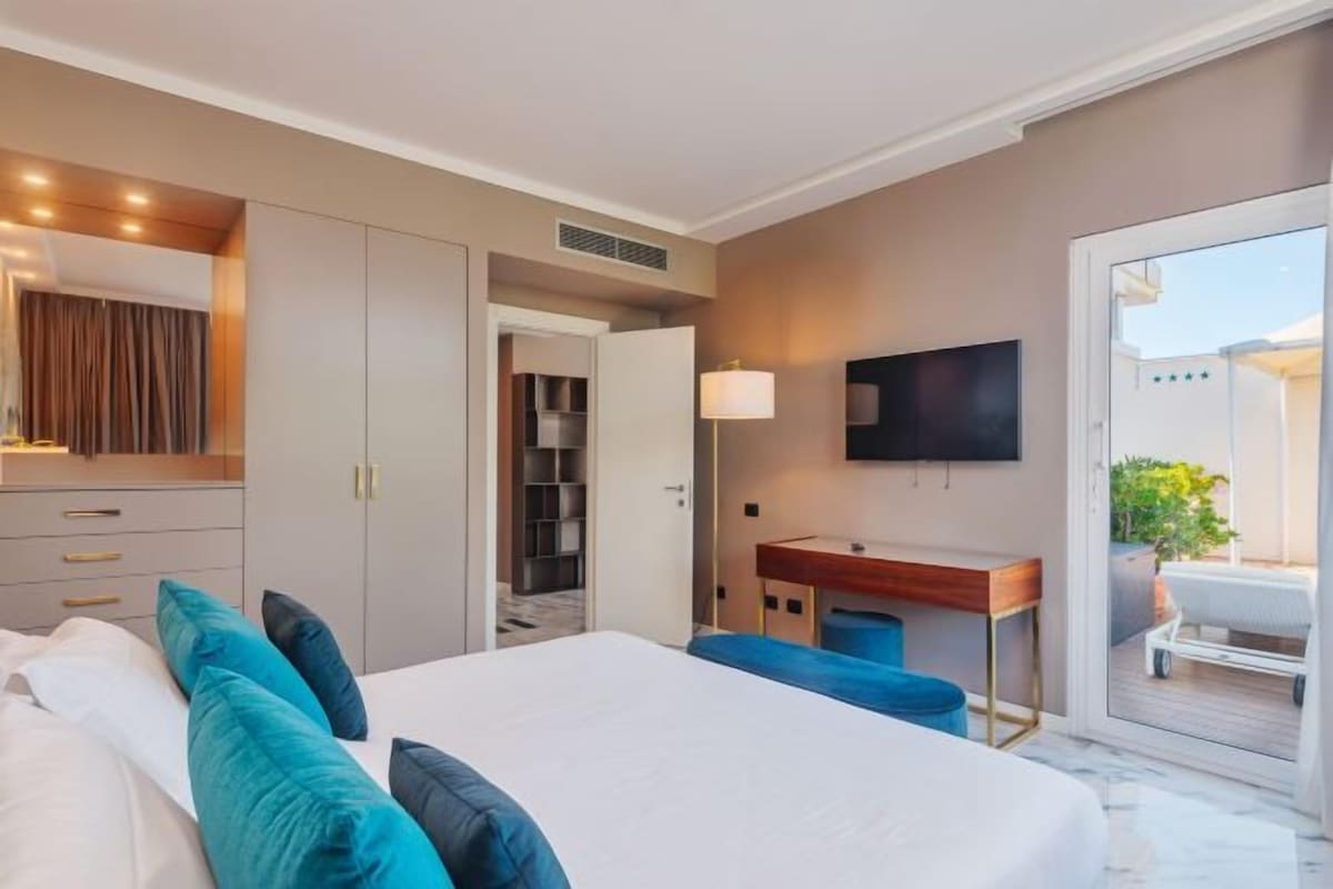A modern hotel room with a large bed, a desk with a chair, a TV, and a sliding door leading to an outdoor area.
