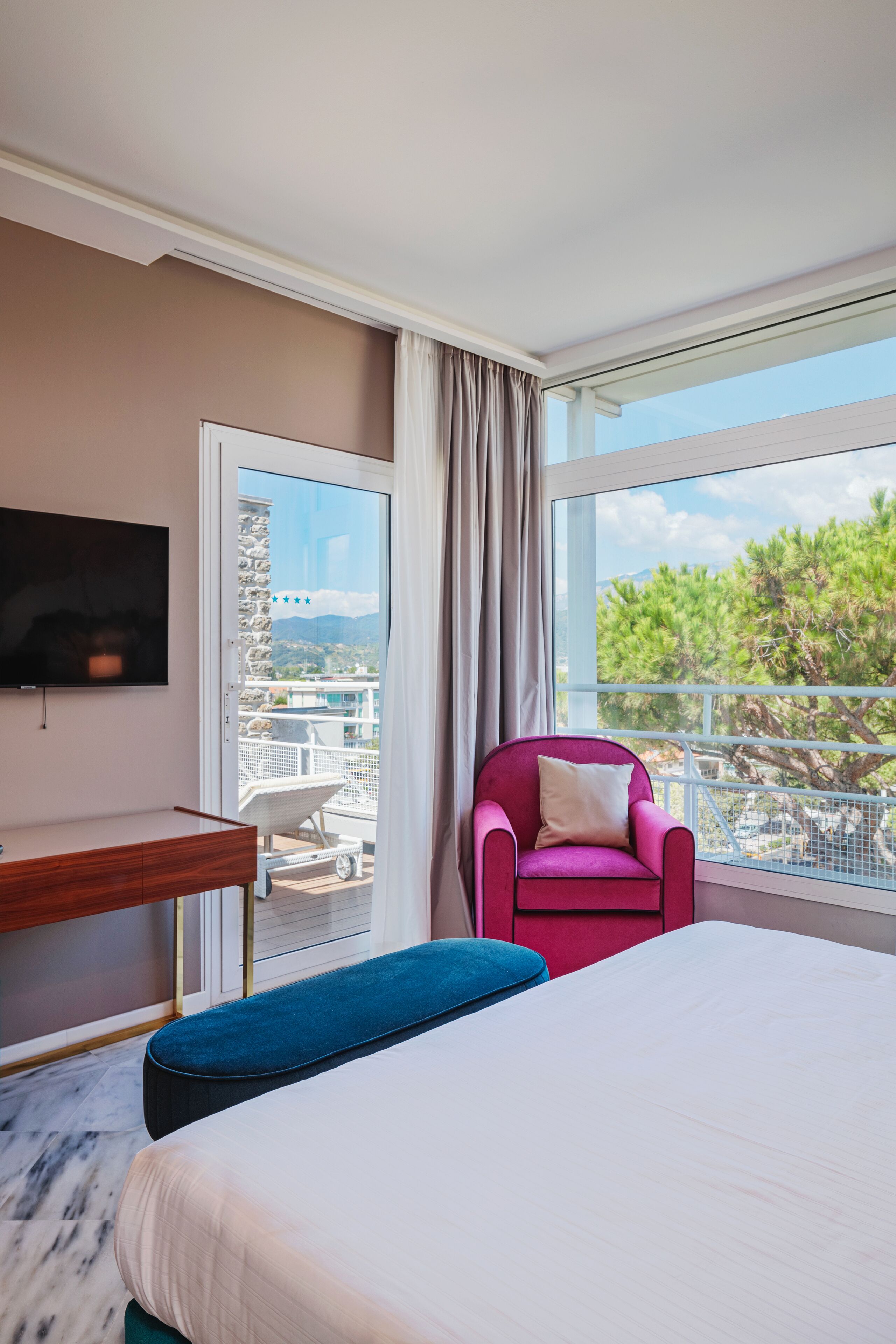 Ein Hotelzimmer mit Bett, Schreibtisch, Fernseher, einem Balkon mit Stuhl und Blick auf Bäume und Berge.