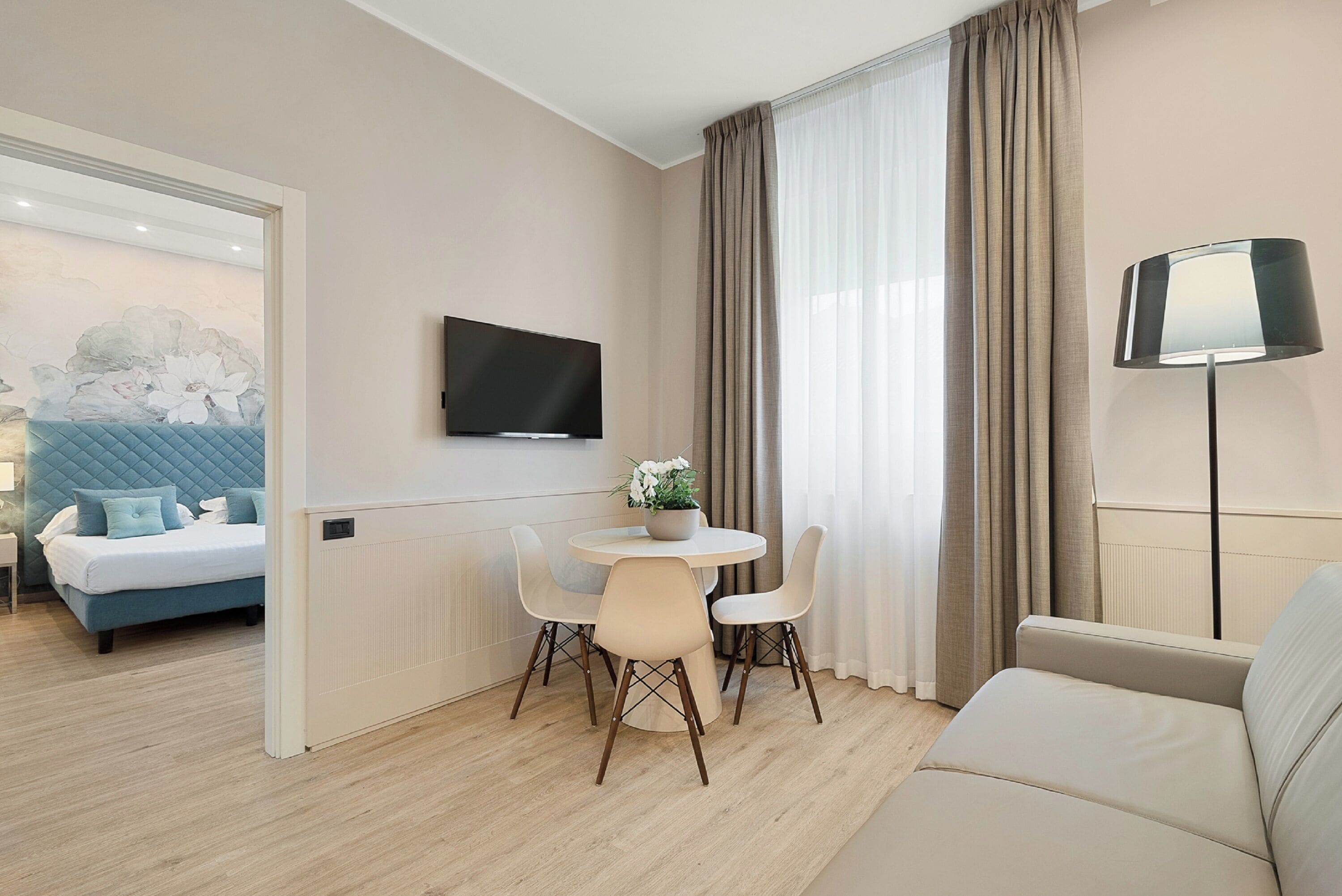 A modern hotel room with a bed, a TV mounted on the wall, a dining area with a table and chairs, and a sofa.