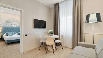 Ein modernes Hotelzimmer mit Bett, Wandfernseher, Essbereich mit Tisch und Stühlen sowie einer Couch.
