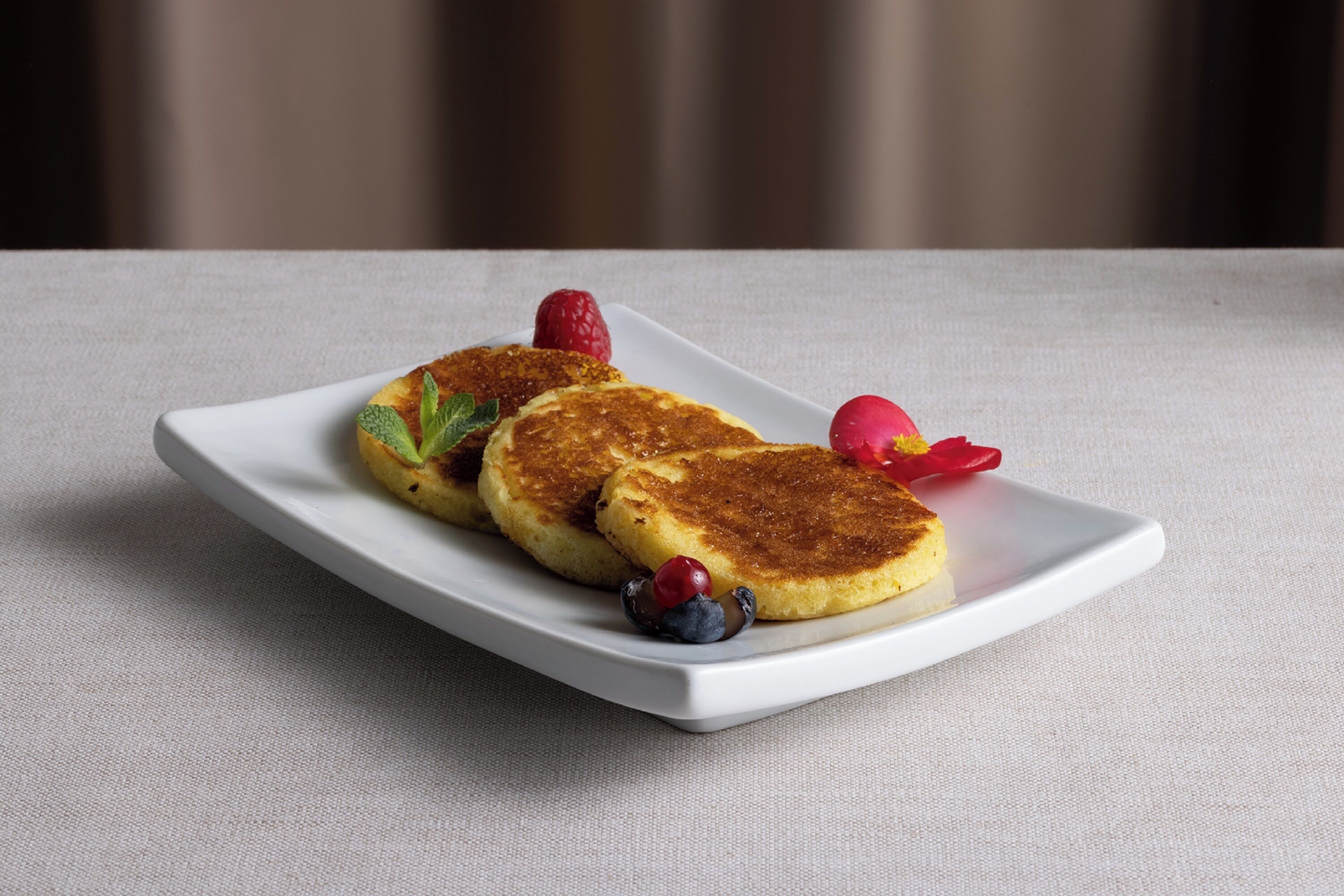 Ein weißer Teller mit drei goldbraunen Pfannkuchen, einer Erdbeere, einem Blaubeere und einer kleinen roten Blüte auf einer strukturierten Oberfläche.