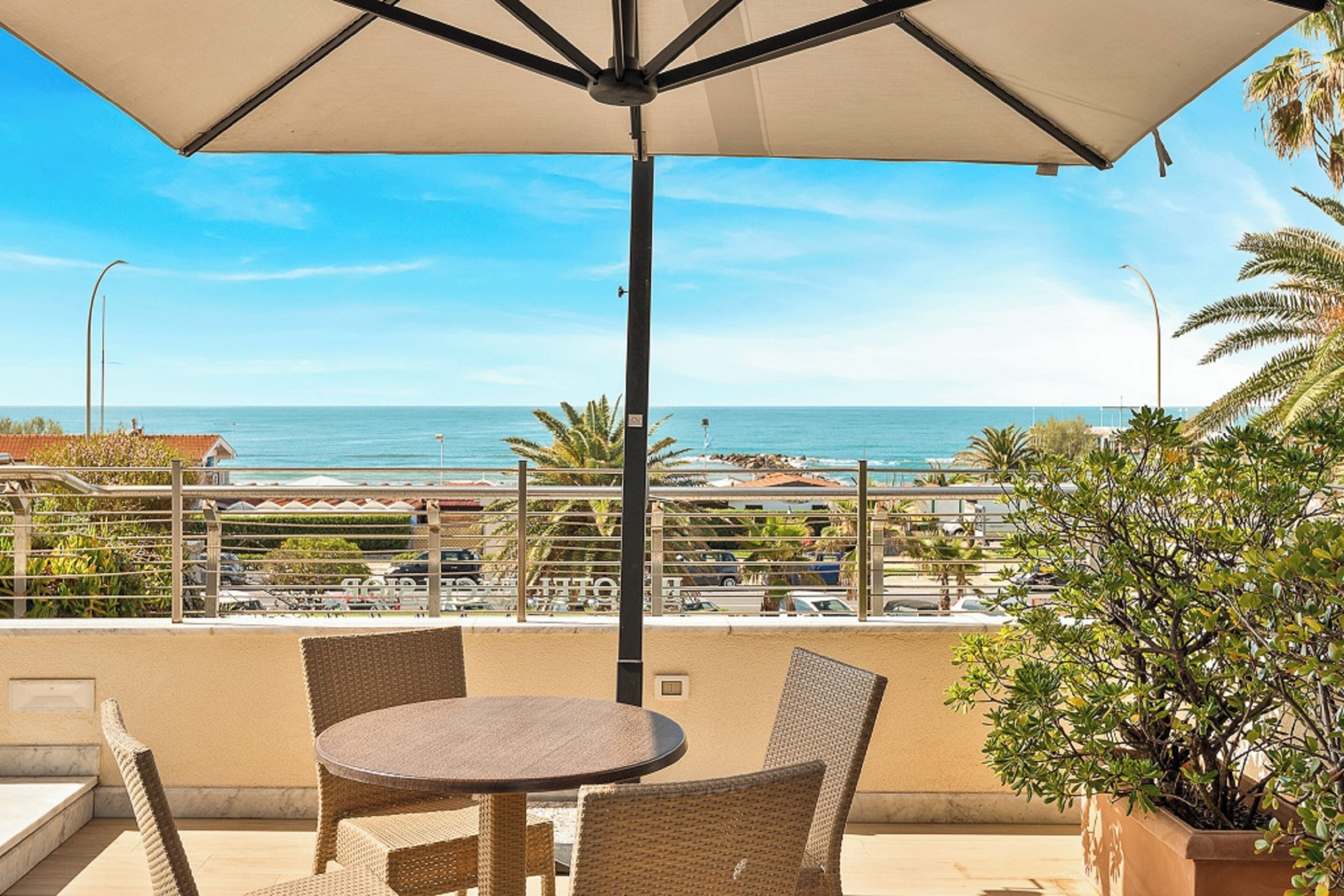 A balcony with a round table and chairs, overlooking a coastal view with the sea and a cityscape.