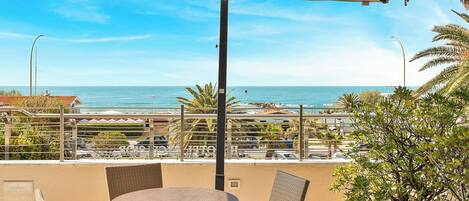 A balcony with a round table and chairs, overlooking a coastal view with the sea and a cityscape.