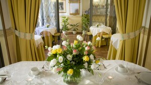Una mesa de comedor dispuesta para una comida, con un florero en el centro, rodeada de sillas y mesas con manteles blancos.