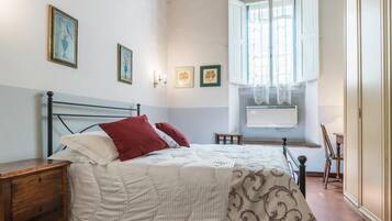 Un dormitorio con techo de madera, una cama con colcha blanca y estampada, una mesita de noche, un ventanal con cortinas de encaje y un radiador fijado a la pared.