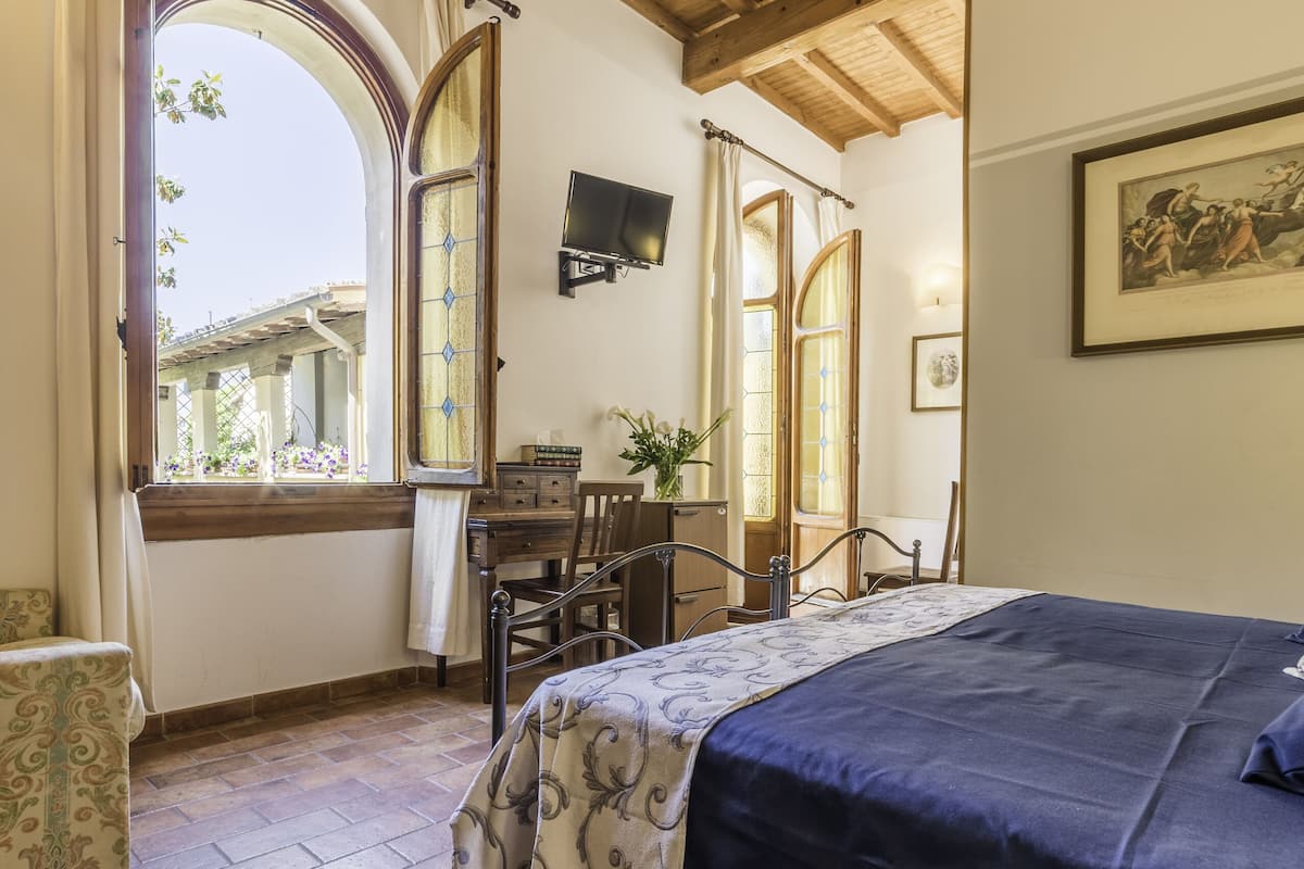 A bedroom with a wooden ceiling, a bed with a blue bedspread, a desk with a chair, a window with curtains, and a painting on the wall.