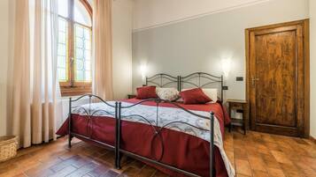 A bedroom with a metal bed frame, a red bedspread, a wooden door, and a window with curtains.