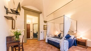 Un dormitorio con cama con dosel, un escritorio con silla y un baño visible a través de un arco.