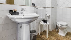 Un baño con lavamanos blanco, espejo y retrete, paredes con azulejos y suelo de baldosas marrones.