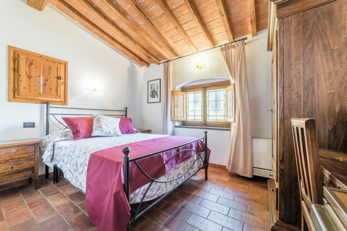 A bedroom with a bed, wooden ceiling beams, a window with curtains, and a wooden wardrobe.