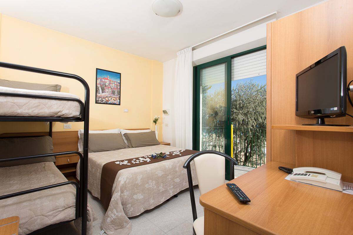 A hotel room with bunk beds, a desk with a telephone, a television, and a window with a view of trees.