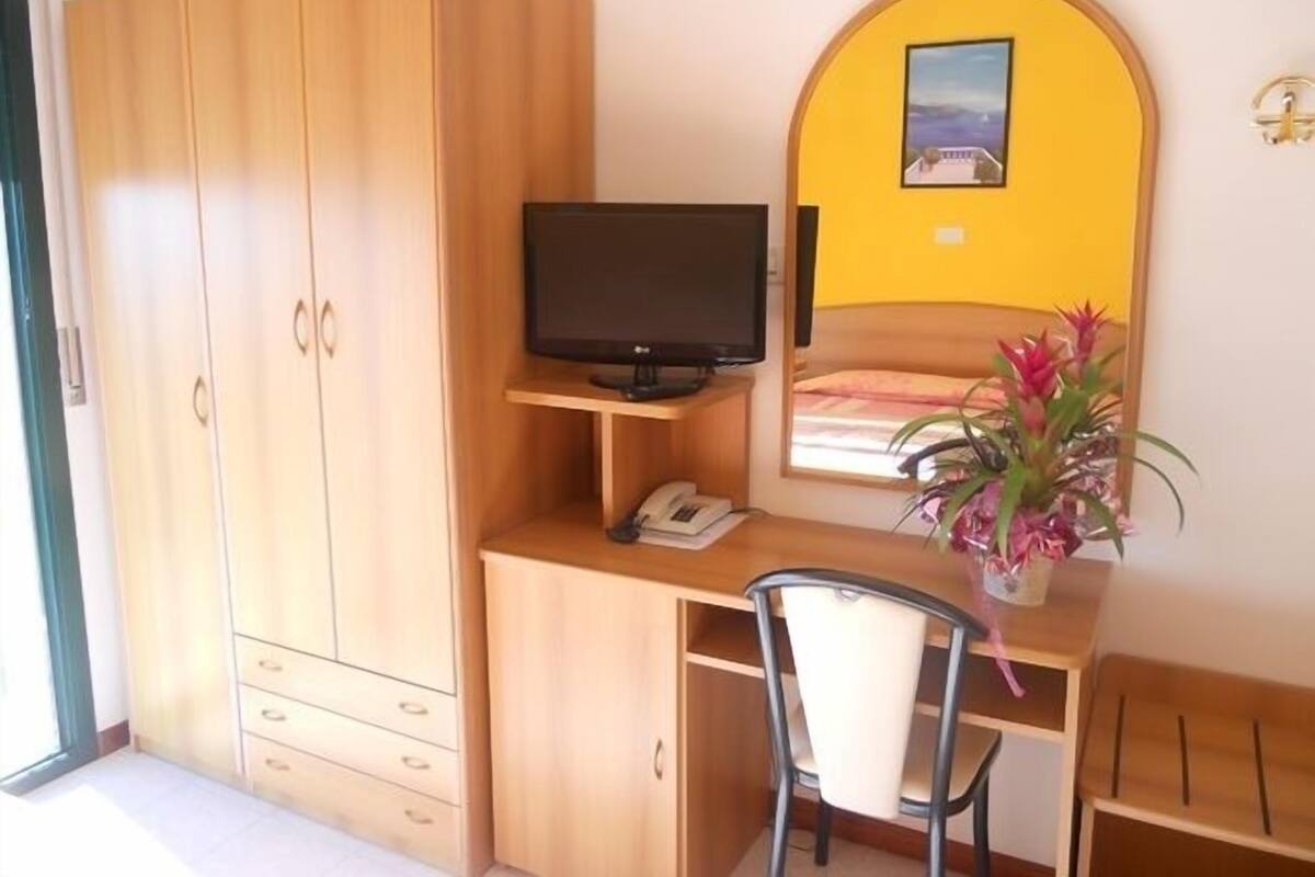 A bedroom with a wooden wardrobe, a desk with a television, a chair, and a bed with a yellow wall and a picture above it.