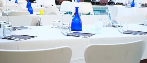 Un salón de banquetes con mesas redondas dispuestas para comer, sillas blancas y una piscina al fondo.