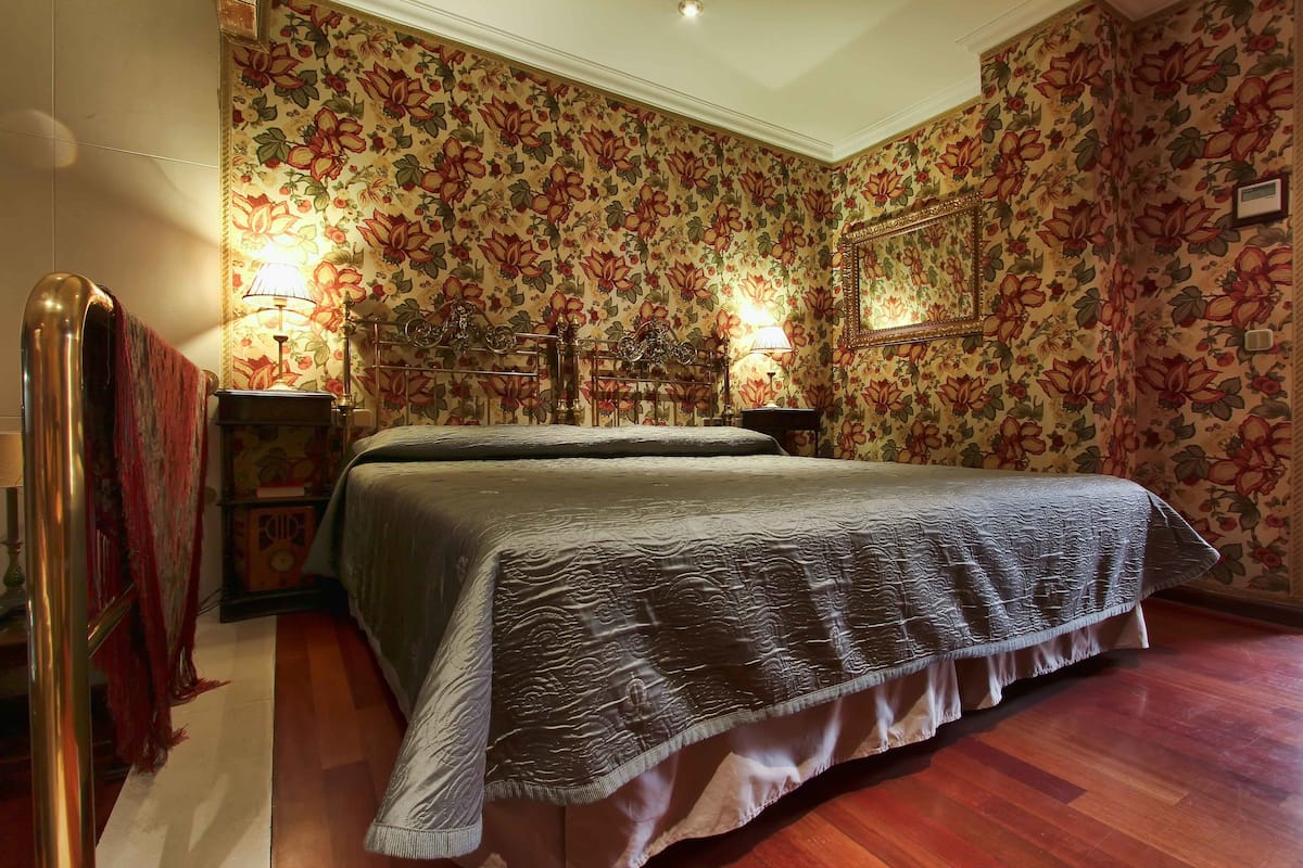 Une chambre avec un papier peint à motifs floraux, un lit recouvert d’une couverture grise, deux lampes de chevet et un plancher en bois.