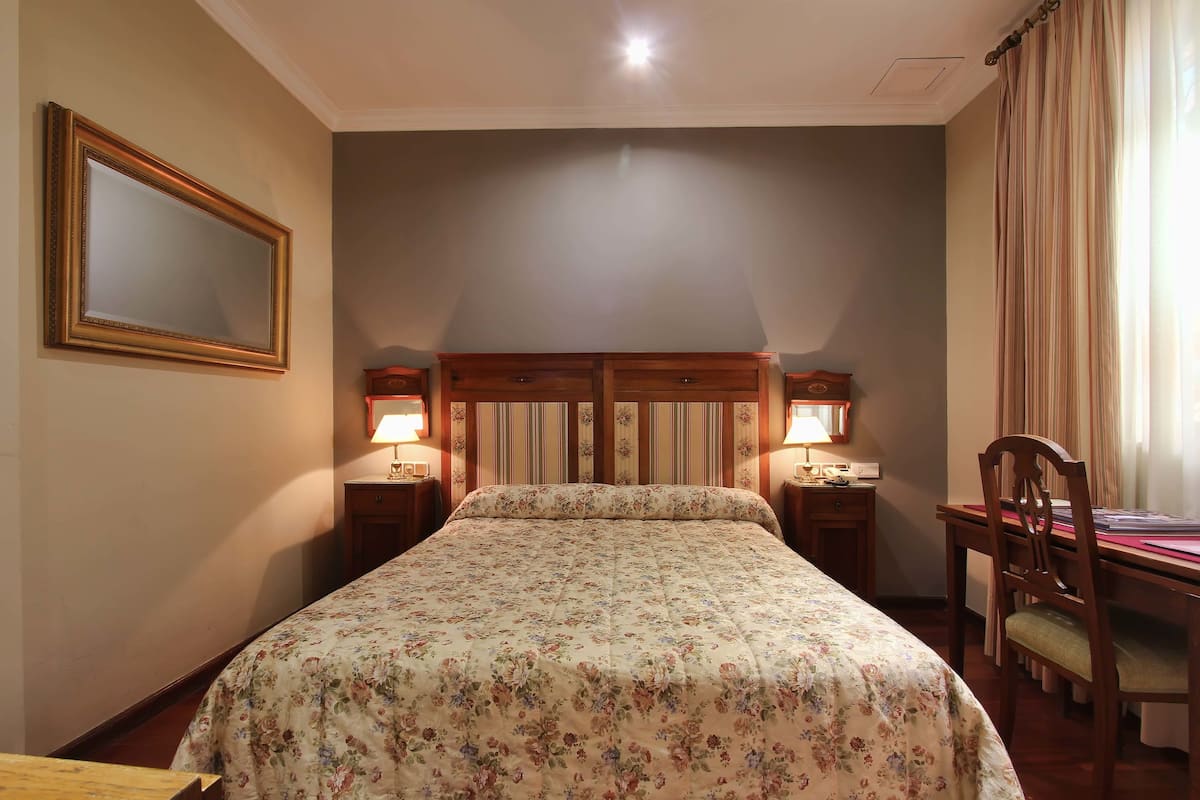 Une chambre d’hôtel avec un lit, deux tables de chevet avec des lampes, un bureau en bois avec une chaise et un grand miroir au mur.