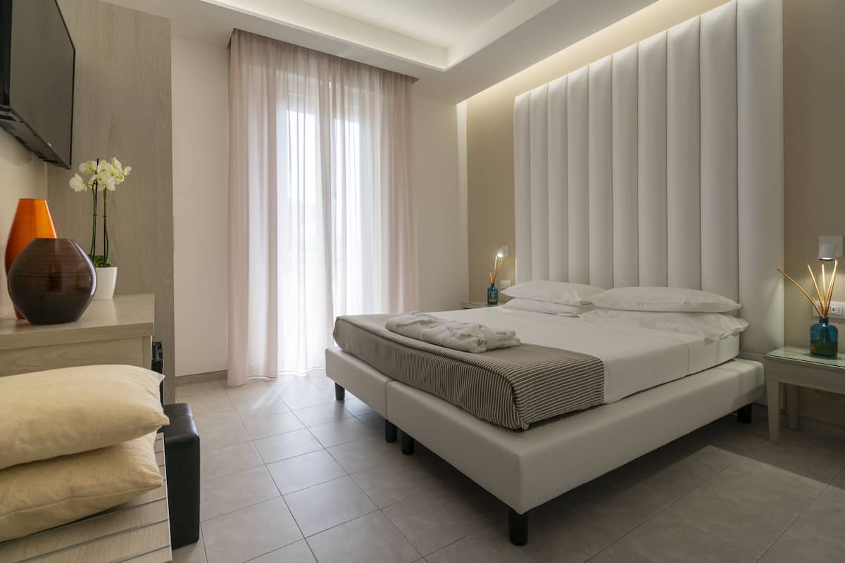 A modern hotel room with a large bed, a bedside table, a nightstand, a TV, and a window with curtains.