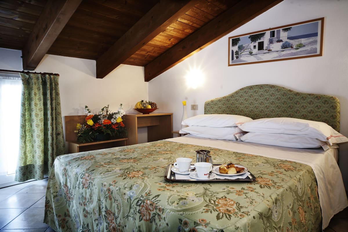 A hotel room with a bed, a nightstand with a flower vase, a tray with breakfast items, and a painting on the wall.