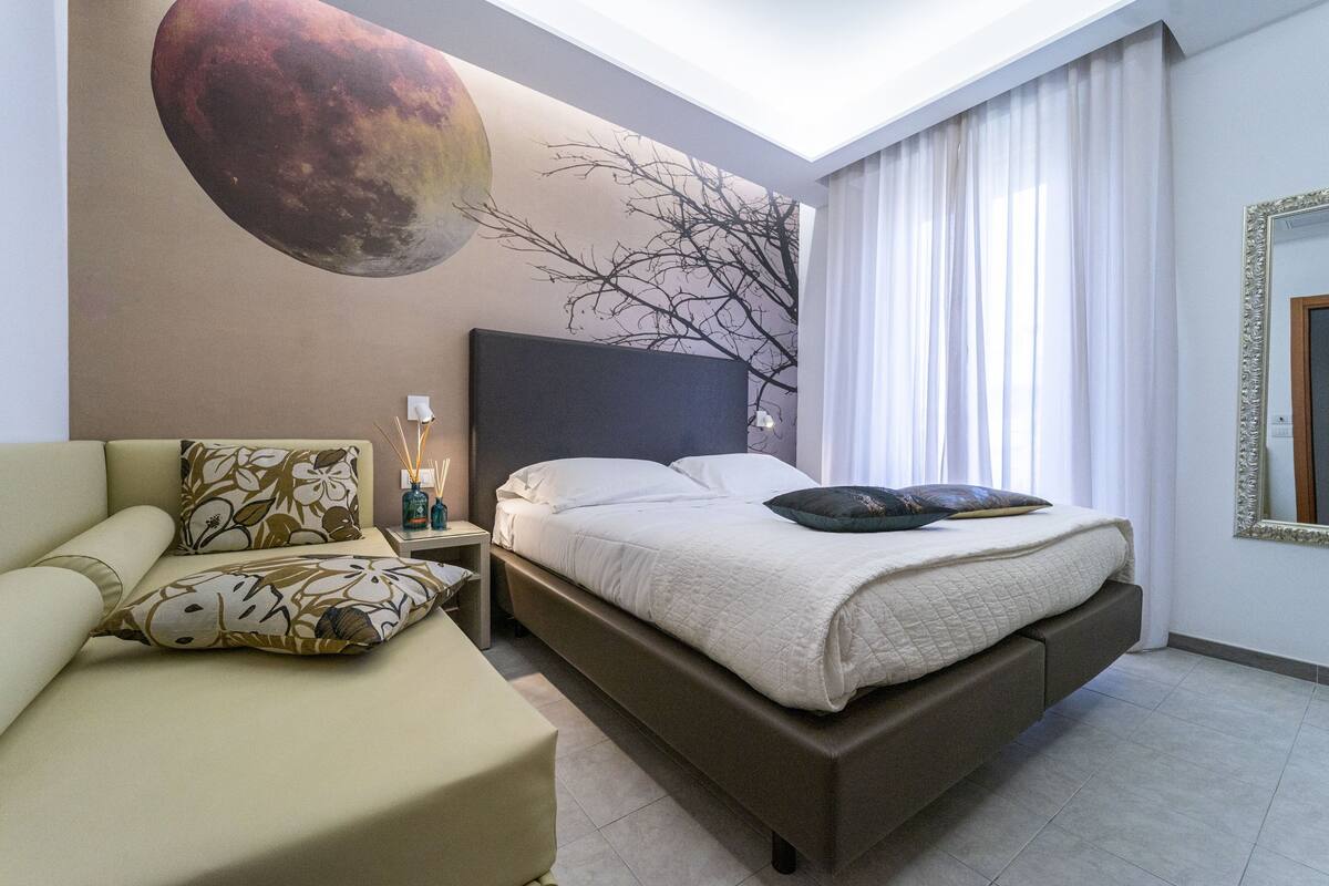 A modern hotel room with a large bed, a sofa, a nightstand with a lamp, and a wall mural.