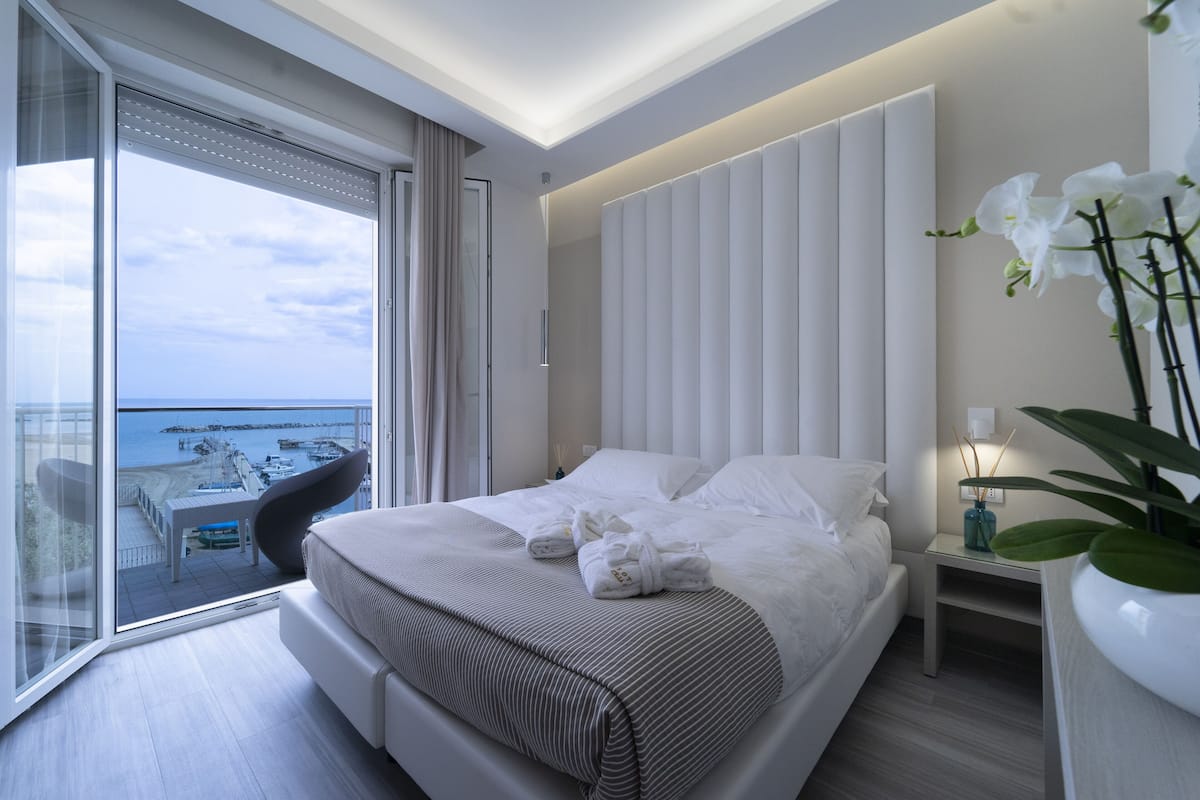 A modern hotel room with a large bed, a balcony with a view of the sea, and a small table with chairs.