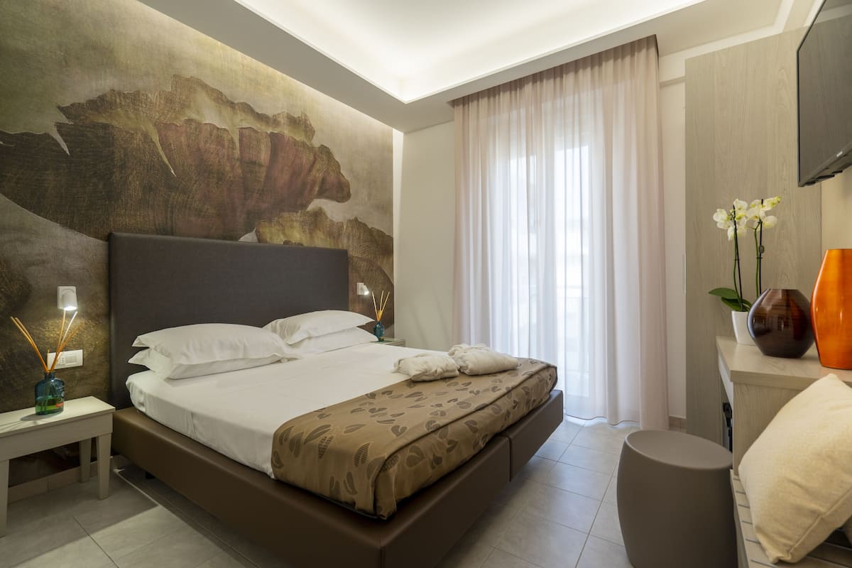 A modern hotel room with a large bed, a nightstand, a bedside table with a vase, and a large window with curtains.