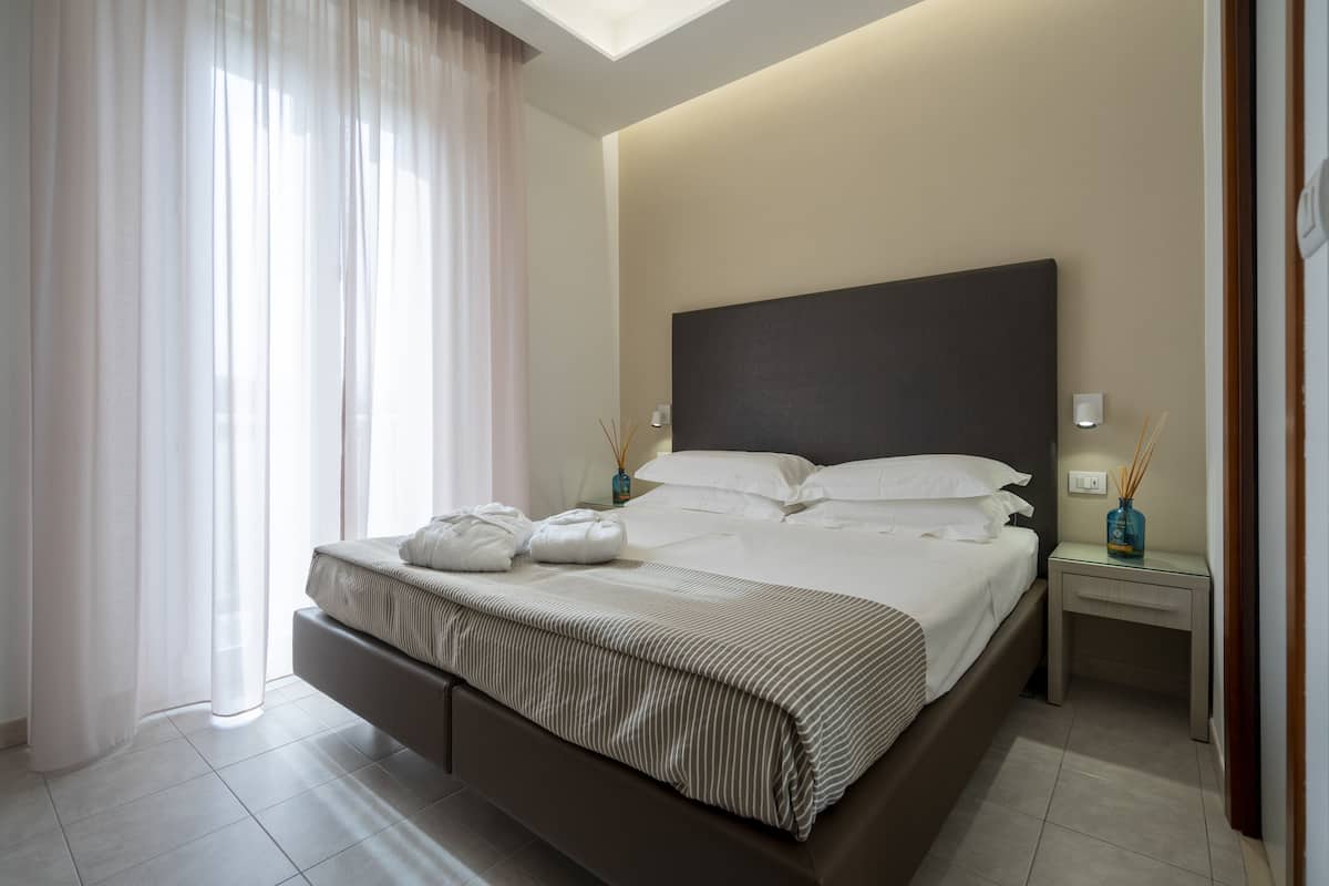 A modern bedroom with a large bed, a nightstand, a bedside table, and a window with sheer curtains.
