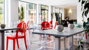 A modern dining area with clear chairs and a mix of red and clear chairs, large windows, and a visible kitchen area. - Hotel AACR Monteolivos (Seville)
