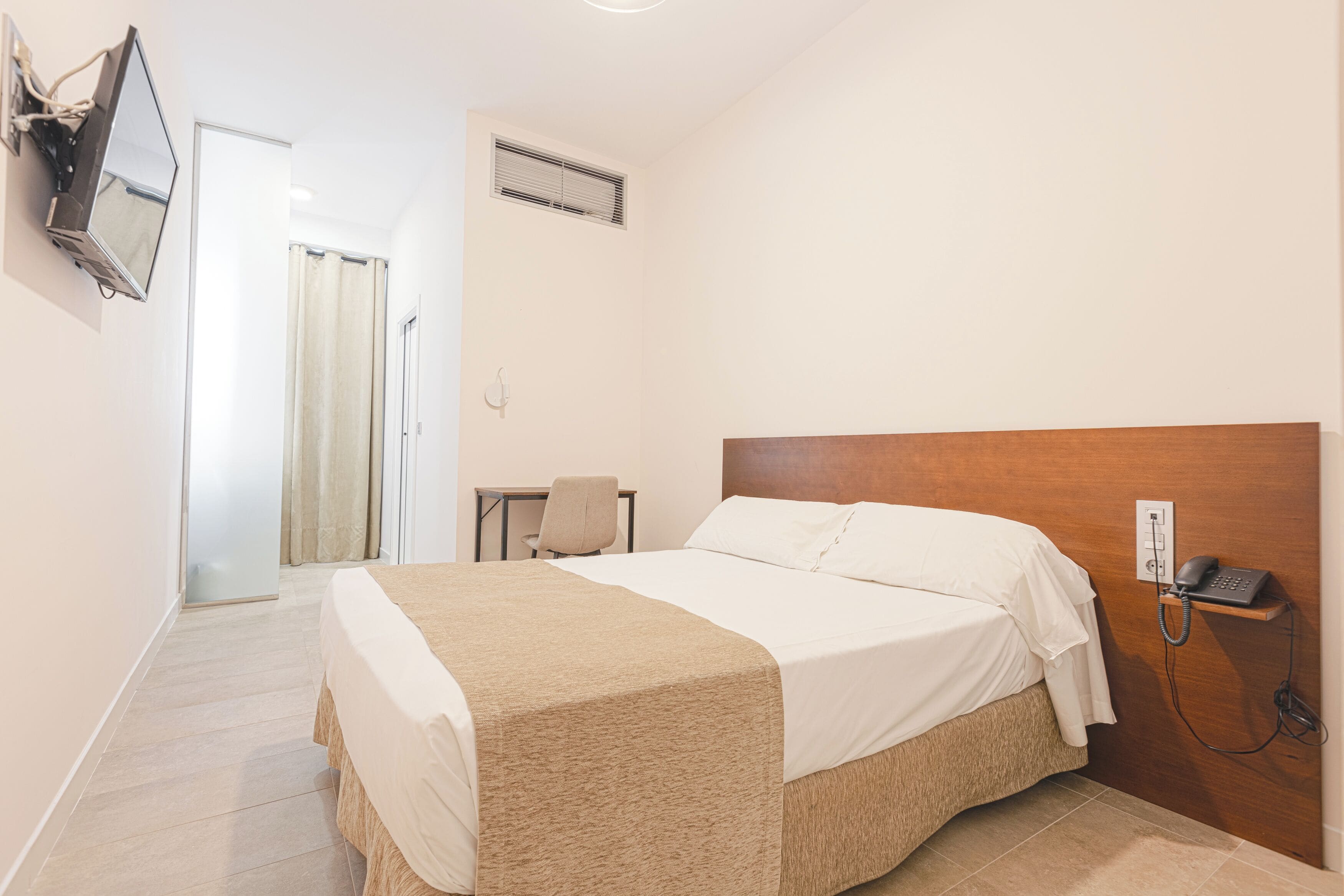 A hotel room with a bed, a desk, a chair, a TV mounted on the wall, and a phone on the wall.