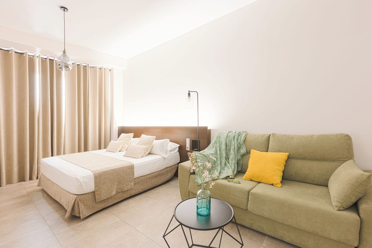 A bedroom with a bed, a green sofa, a small table with a vase, and a lamp.