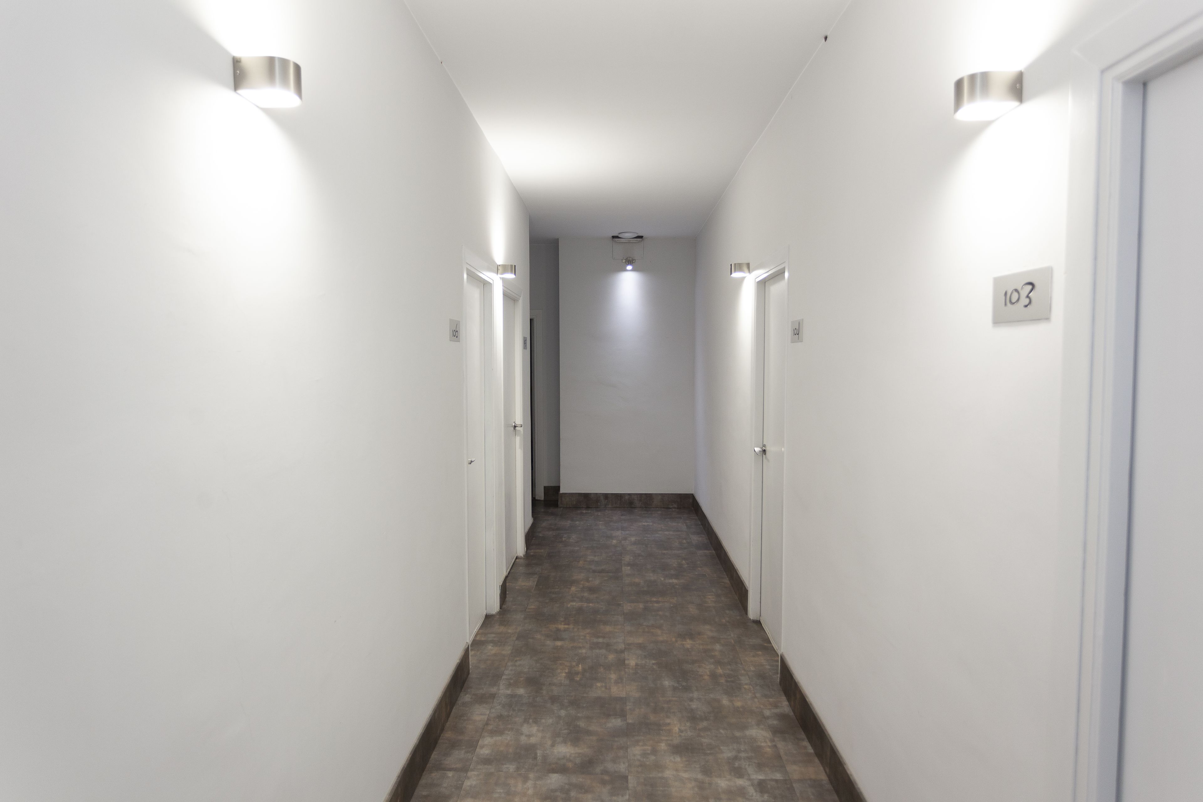 Un pasillo estrecho con paredes blancas y suelo de baldosas, que conduce a una puerta con el número 103.