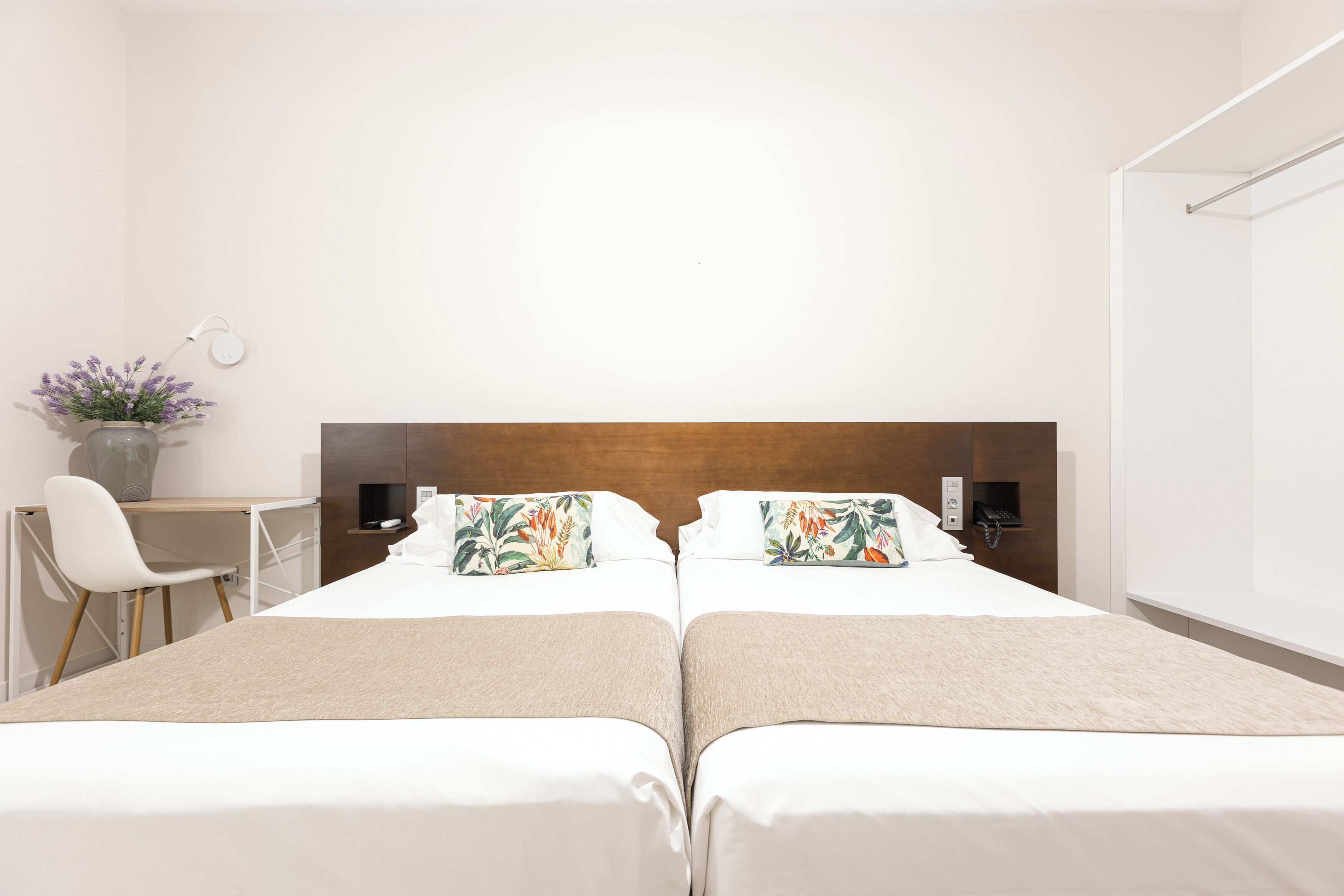 A hotel room with two beds, a desk, a chair, a vase with flowers, and a wall-mounted light.