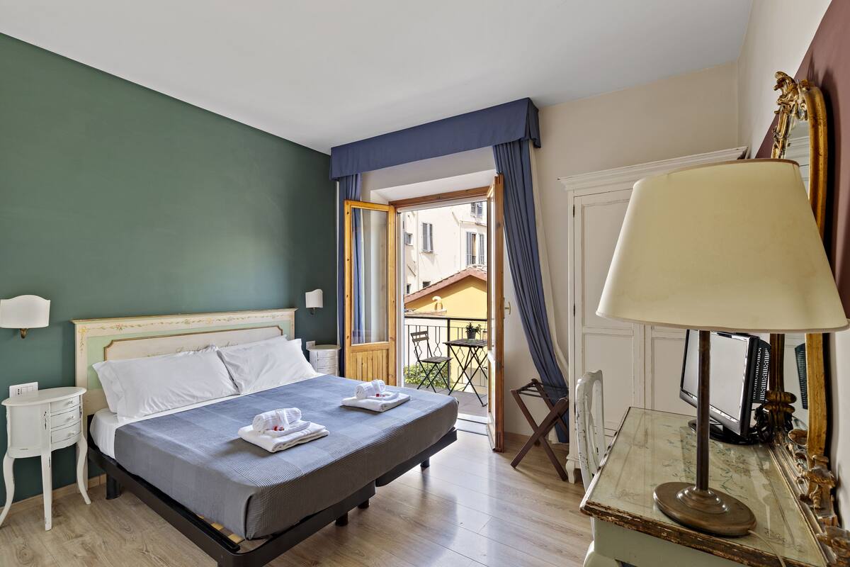 Un dormitorio con cama, mesita de noche, lámpara y vistas al balcón exterior.
