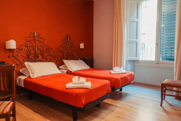A hotel room with two beds, each with a red bedspread and white pillows. There is a wooden chair and a small table with a lamp.
