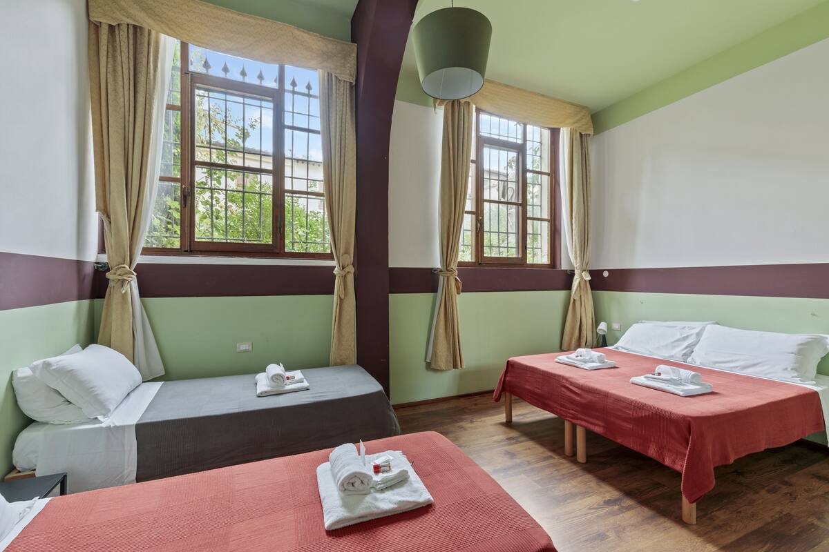 Una habitación con dos camas, cada una con cubrecama rojo y almohadas blancas, una mesita con mantel rojo y una ventana con cortinas.