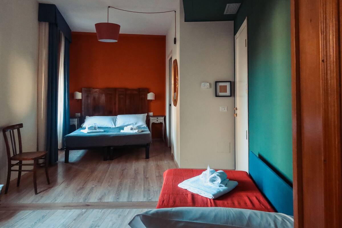 Habitación de hotel con piso de madera, cama con colcha roja, una silla de madera y lámpara colgante.