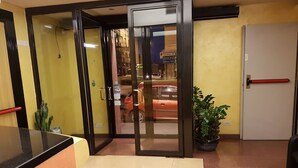 Un hall d’hôtel avec une entrée dotée d’une porte en verre, une plante en pot, une poignée de porte rouge et un sol carrelé.
