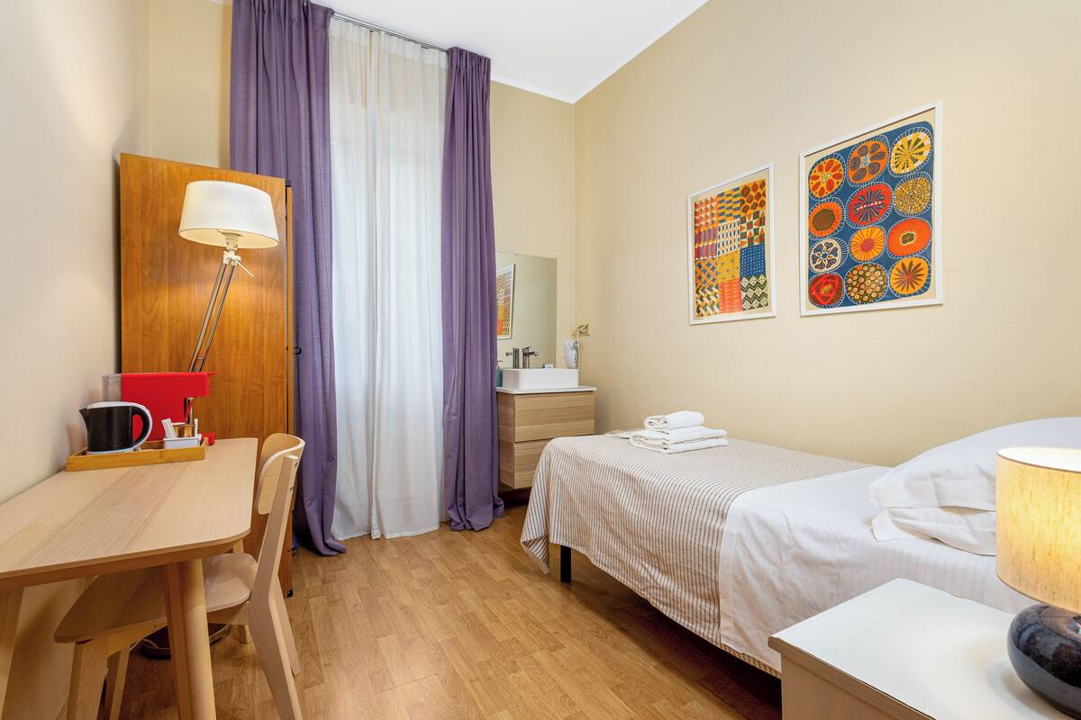Una camera d'albergo con un letto, un comodino, una scrivania con sedia e uno specchio.