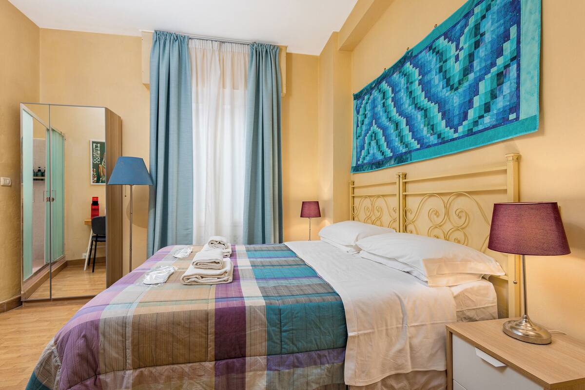 Una camera d'albergo con un letto, un comodino, una lampada, uno specchio, una finestra con tende e un quadro appeso al muro.