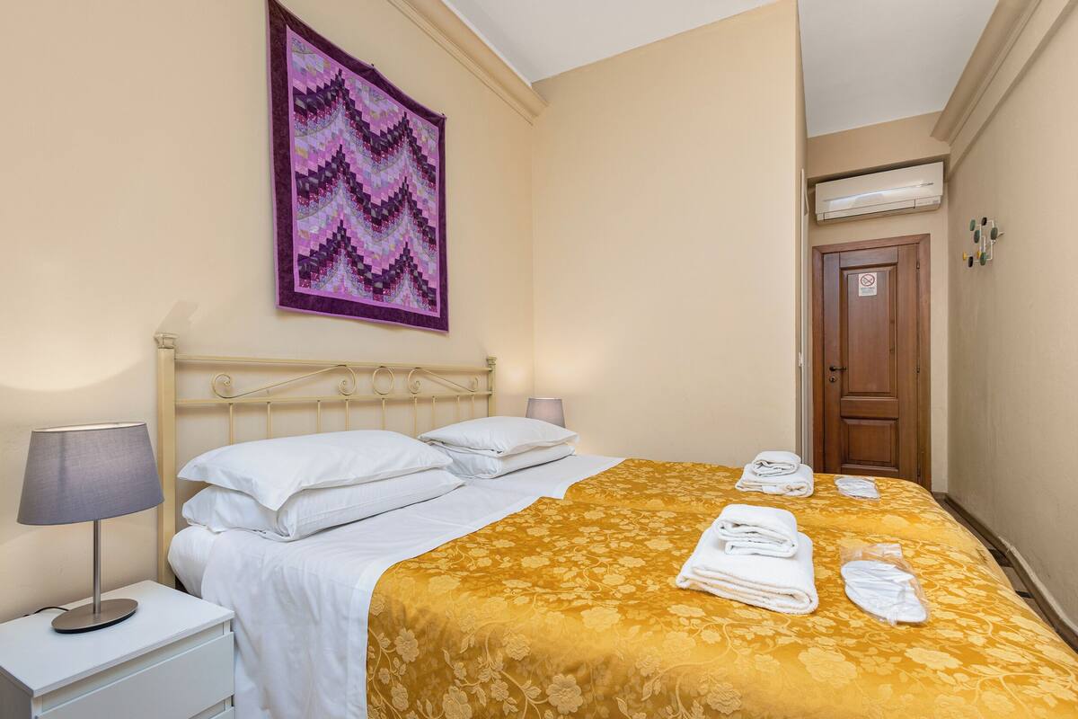 Camera d'albergo con un letto, un comodino, una lampada, un elemento decorativo a parete e una porta con un cartello.