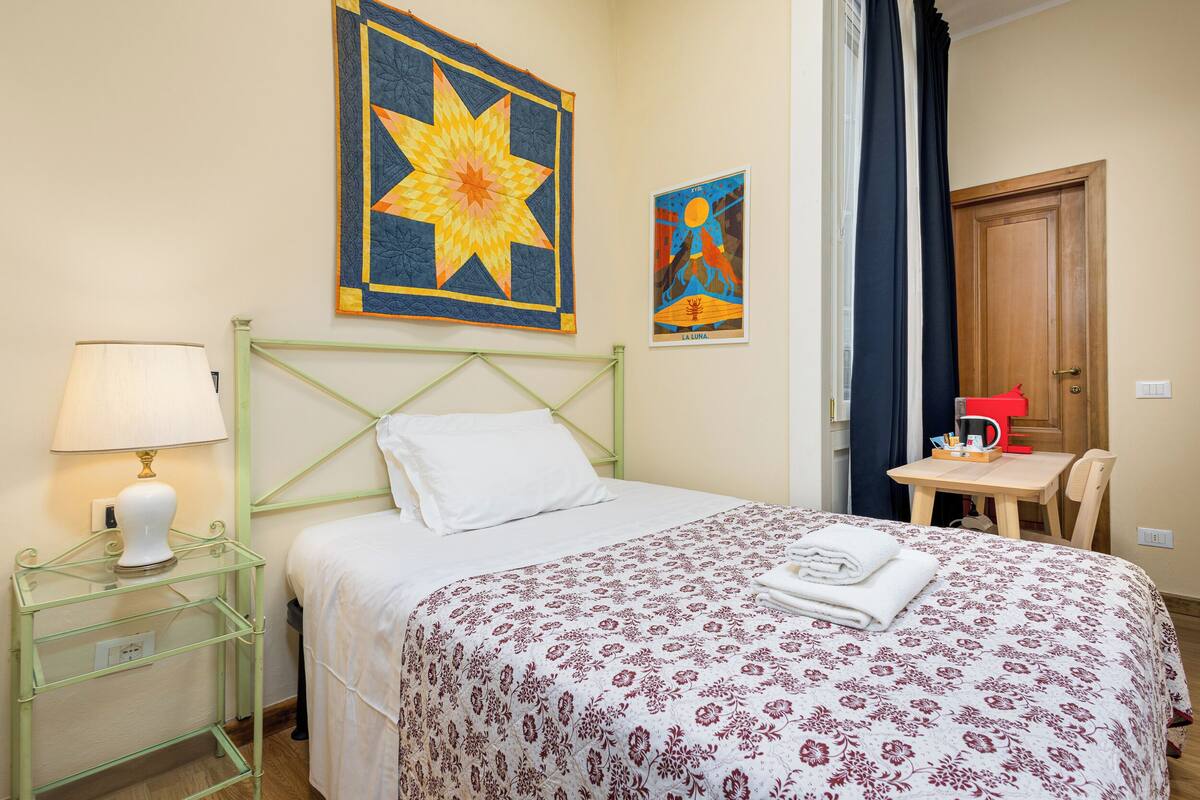 Una camera d'albergo con un letto, un comodino con una lampada, un tavolino con una lampada rossa e una porta.