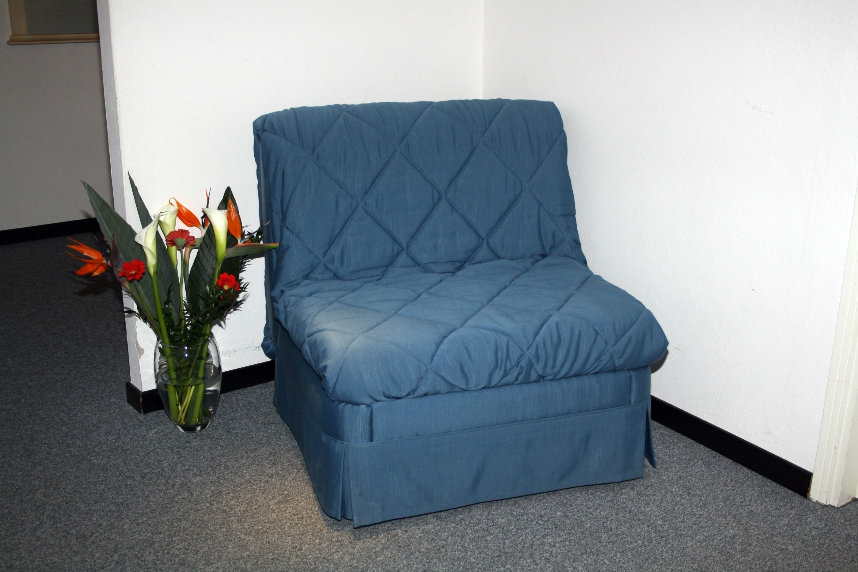 A blue sofa in a corner of a room with a white wall and a grey carpet.