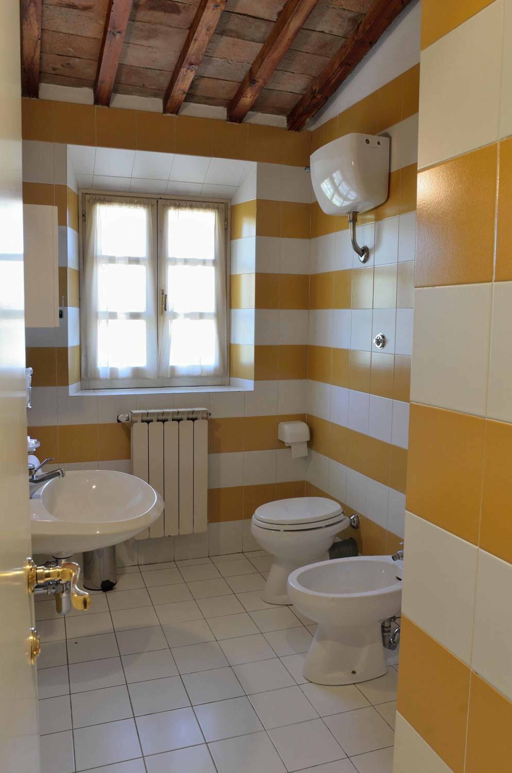 A bathroom with striped yellow and white tiles, a window, a radiator, a sink, a toilet, and a bidet.