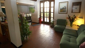 Un salón con un sofá verde, una mesita con una lámpara y una planta en maceta. Se ve una puerta que da a una zona de cocina con una ventana.