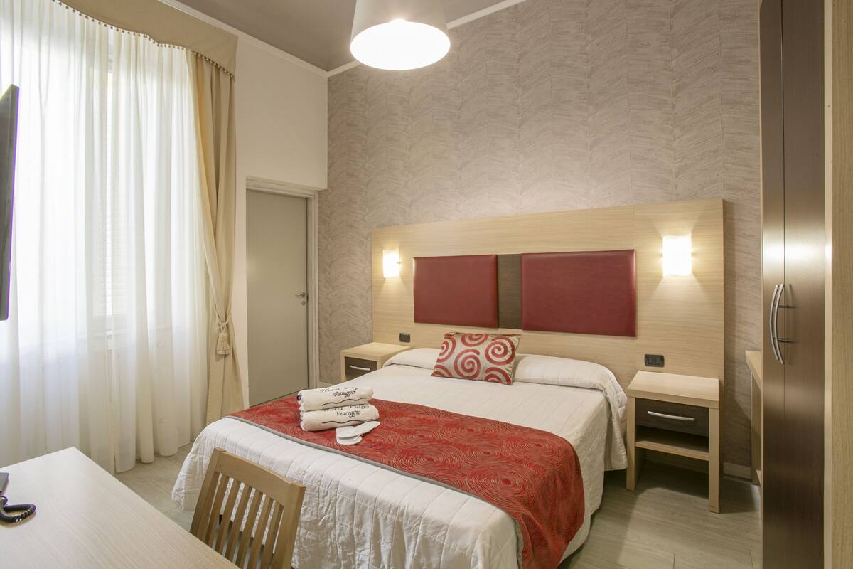A hotel room with a large bed, two bedside tables, a wooden wardrobe, a desk, and a chair.