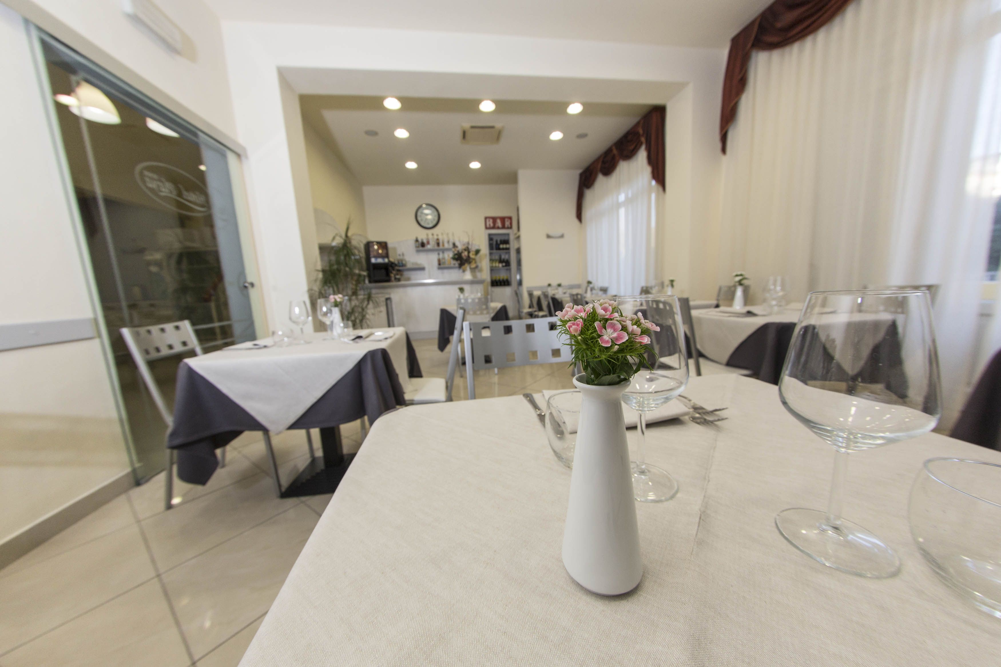 A dining area with tables set for two, a white vase with pink flowers, and a kitchen in the background.