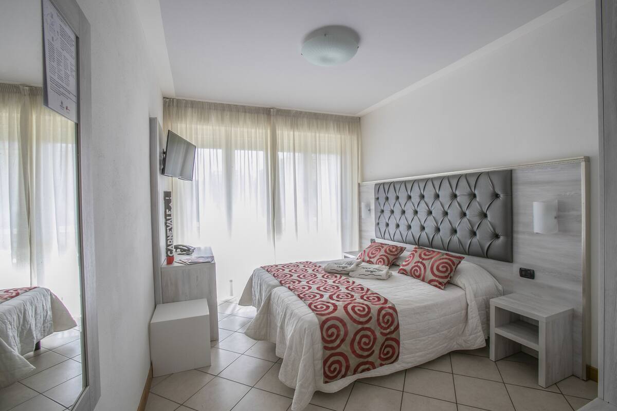 A modern hotel room with a large bed, a bedside table, a TV mounted on the wall, and a window with sheer curtains.