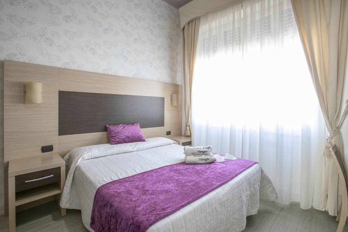 A hotel room with a bed, a nightstand, a headboard, and a window with curtains.