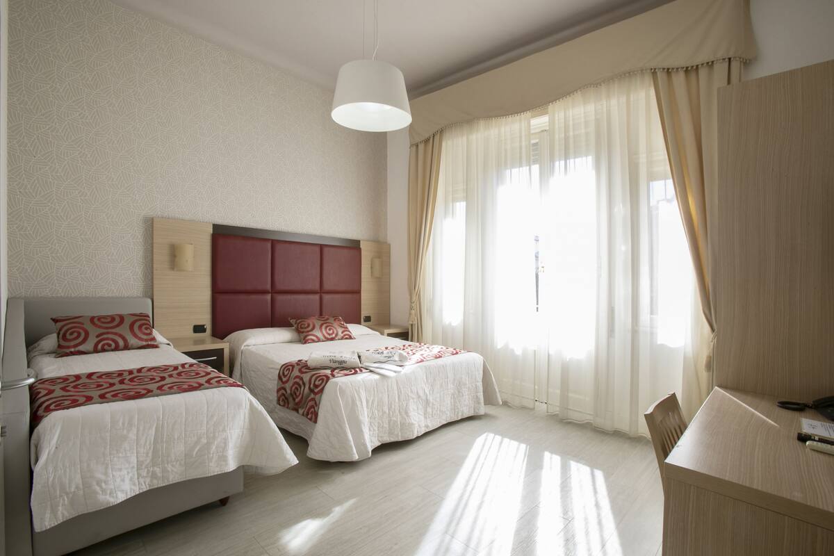 A hotel room with two beds, a desk, and a large window with sheer curtains.